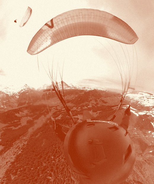 Pilot's view from under a paraglider wing, with a friend's wing in the corner and an alpine valley below.